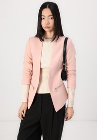 Light pink blazer with a fitted silhouette, two zippered pockets, rolled-up sleeves, paired with a cream ribbed top and black pants.