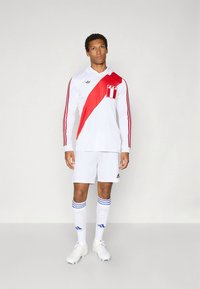 Άσπρη ποδοσφαιρική φανέλα μακρυμάνικη με κόκκινη διαγώνια ρίγα, που φέρει ένα έμβλημα και τρεις μαύρες ρίγες στα μανίκια· σετ με άσπρα σορτς και κάλτσες.