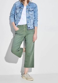 Chaqueta de mezclilla azul claro con dos bolsillos en el pecho, camiseta blanca, pantalones anchos verdes y zapatillas beige con acentos texturizados.