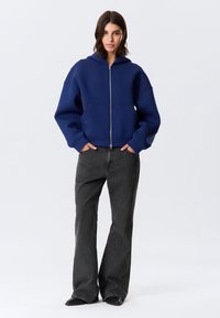 Μπλε σκούρο hoodie με φερμουάρ και κουκούλα, μεγάλες μπροστινές τσέπες και φαρδιά εφαρμογή, συνδυασμένο με σκούρο γκρι φαρδιές τζιν παντελόνες και μαύρα παπούτσια.