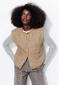 Gilet en laine beige avec un col rond, des poches avant et des boutons dorés, associé à une chemise à rayures à manches longues et un pantalon à carreaux.