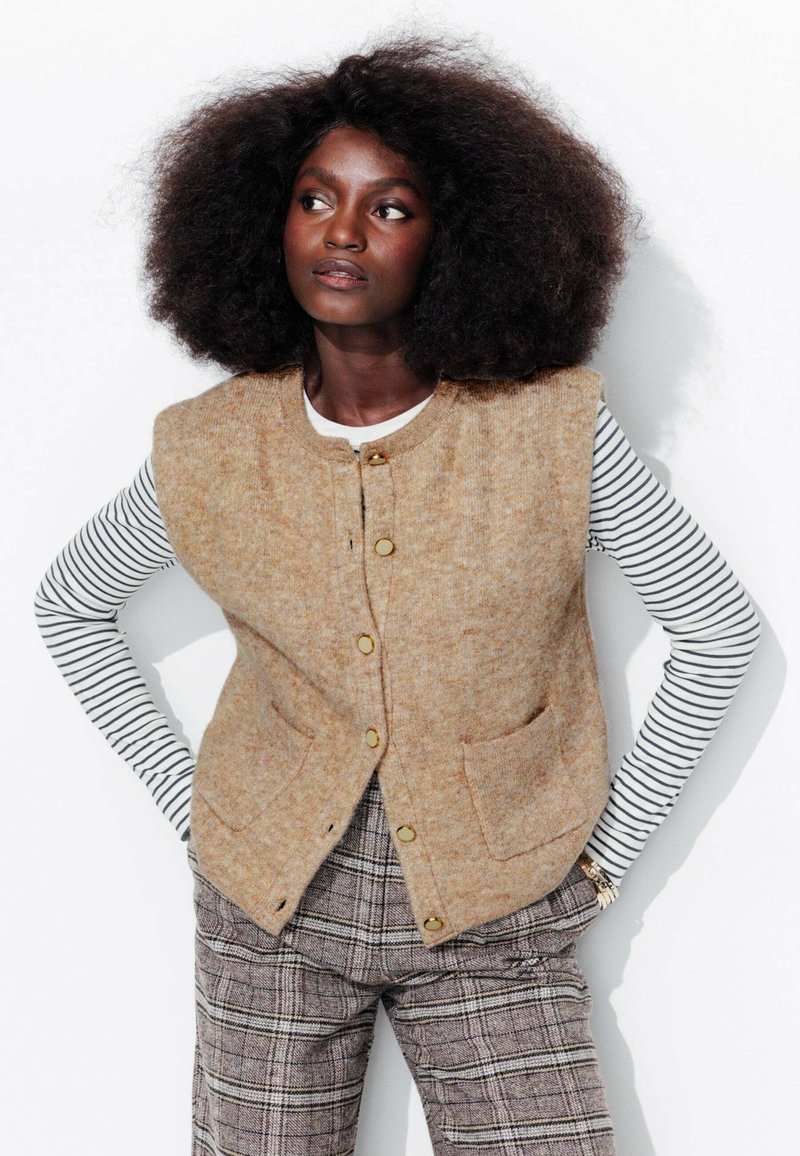 Gilet en laine beige avec un col rond, des poches avant et des boutons dorés, associé à une chemise à rayures à manches longues et un pantalon à carreaux.