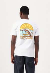 Bijela pamučna majica s živopisnim grafičkim prikazom surfer na valovima, s dizajnom žutog sunčevog sjaja i natpisom "Rip Curl."