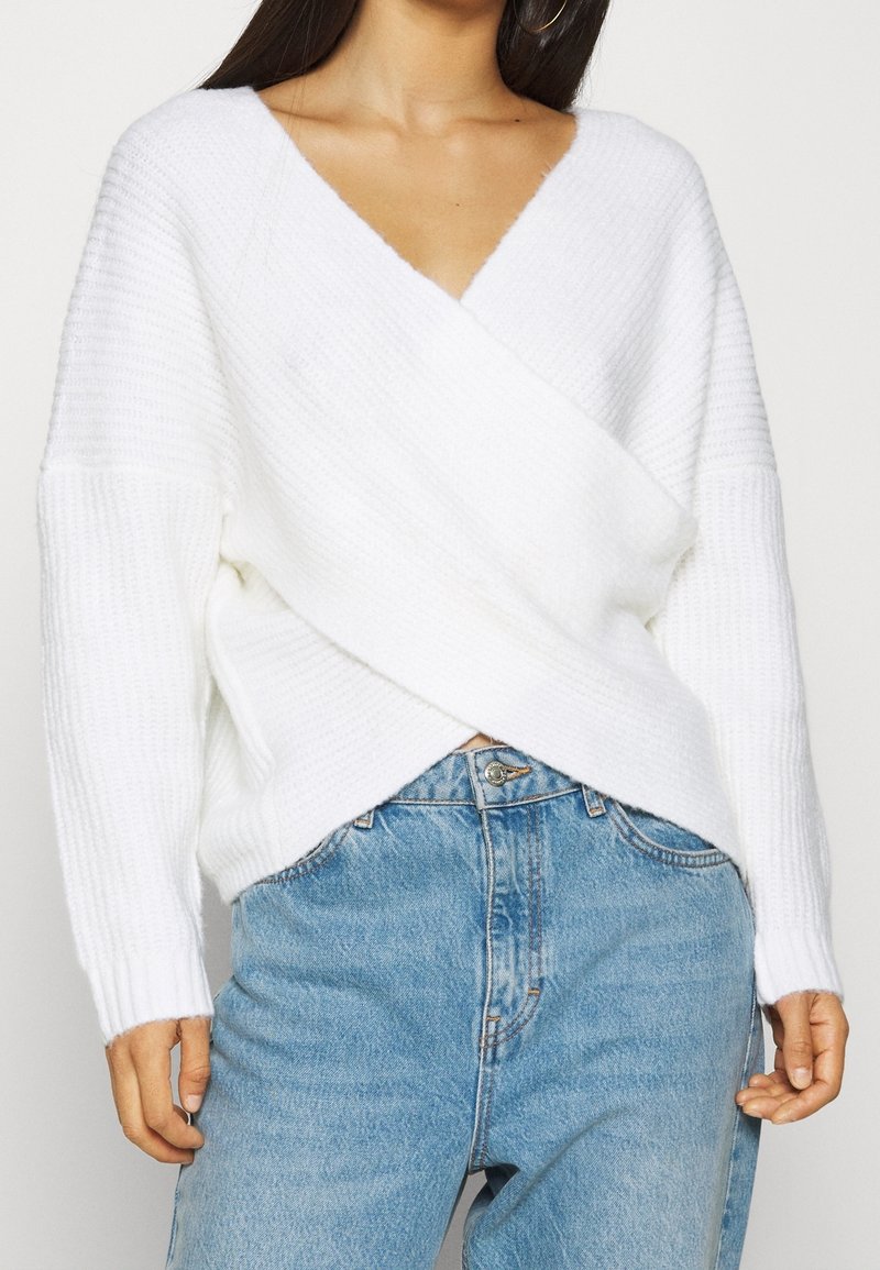 Weißer gestrickter Wickelpullover mit tiefem V-Ausschnitt, strukturiertem Stoff und langen Ärmeln, kombiniert mit hoch taillierten blauen Jeans.