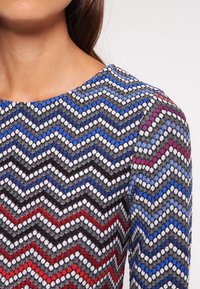 Svetr s vzorem v designu cik-cak v modré, červené, černé a bílé barvě, s kulatým výstřihem a texturovanou tkaninou.