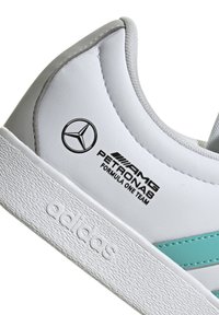 Vit sneaker med grå accentfärger, med teal-ränder och texturerad gummisula. Visar Mercedes-Benz och AMG Petronas varumärke.