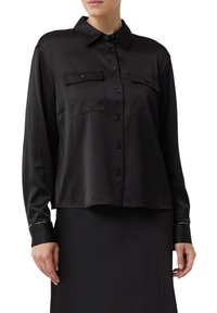 Woman wearing a black long-sleeve button-up shirt with two chest pockets and a black skirt against a white background.