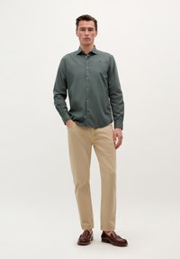 Camisa de botón de manga larga, de color verde oscuro y corte ajustado, combinada con chinos beige y mocasines marrones, contra un fondo neutro.