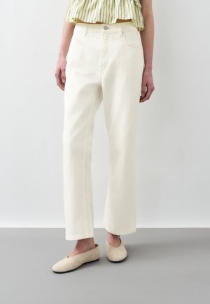 Személy törtfehér egyenes szárú nadrágot, bézs bebújós cipőt és rövidített, sárga-zöld csíkos felsőt visel, és szürke padlón áll.