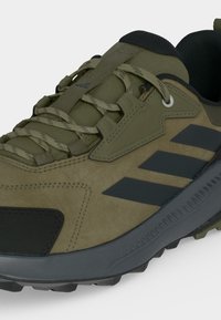 Chaussure de randonnée vert olive avec des rayures noires, col rembourré, semelle épaisse texturée et lacets à motifs sur fond gris clair.