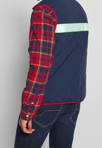 Marinblå quiltad väst med en reflekterande grön och vit rand, buren över en röd och marinblå rutig skjorta, i kombination med mörka jeans.