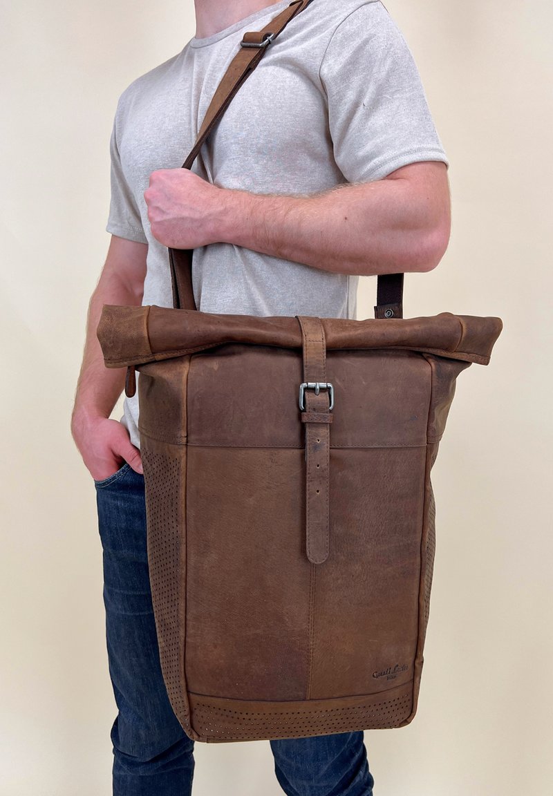 Bruine leren roll-top tas met verstelbare schouderriem, metalen gesp en geperforeerde accenten. Gelaagde oppervlakte met een gestructureerd ontwerp.