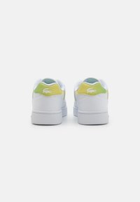 Sneakers bianchi con accenti gialli e verdi sul retro, materiale in pelle liscia e suola testurizzata con un piccolo logo vicino al tallone.