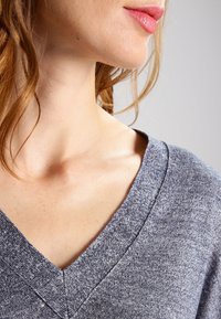 Šedý top s véčkovým výstřihem, vyrobený z jemné, strukturované látky. Výstřih je žebrovaný, s nepravidelným vzorem a uvolněným střihem kolem límce.
