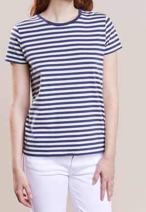 Kobieta ubrana w granatowo-biały t-shirt w poziome paski z krótkim rękawem i białe spodnie na jednolitym tle.