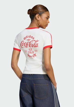 Kvinde med knold, iført hvid Adidas- og Coca-Cola-t-shirt med rød kant, blå jeans med sideløb, set bagfra.
