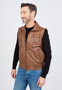 Braune, ärmellose Lederjacke mit Reißverschluss, mehreren Fronttaschen, geripptem Saum und glatter Textur. Getragen über einem schwarzen Langarmshirt.
