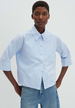 Persoon draagt een lichtblauw overhemd met driekwart mouwen en blauwe spijkerbroek, staat met de handen in de zij.