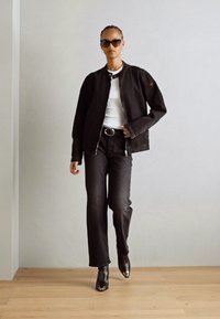 Giacca nera con zip, top bianco, jeans neri e stivali con tacco neri; la modella indossa occhiali da sole; sfondo neutro.