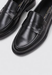 Mocasines de cuero negro con punta redonda, que presentan un diseño cosido y un detalle de doble hebilla en la parte frontal. Textura suave, tacón bajo.