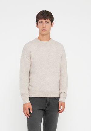 Lichtbeige gebreide trui met een ronde hals, geribde manchetten en zoom; gecombineerd met grijze jeans, tegen een effen achtergrond.
