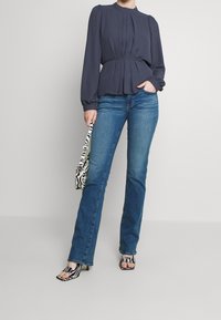 Marinebluse mit Falten, kombiniert mit blauen Denim-Jeans. Die Accessoires umfassen eine Tasche mit Zebramuster und offene Sandalen mit Blockabsatz.