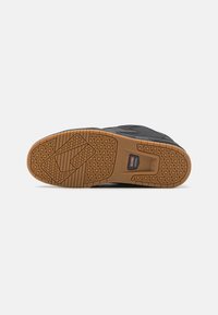 Black sneaker with a textured upper, featuring a brown rubber sole. The sole has a geometric pattern and a branded detail in the center.