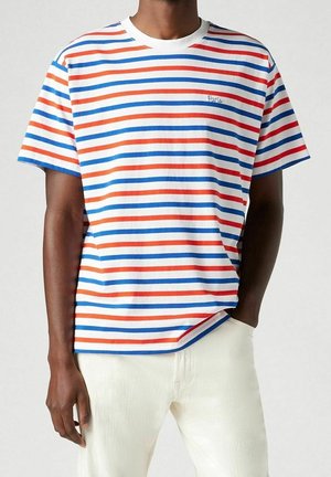 Homme portant un t-shirt à manches courtes avec des rayures horizontales rouges, bleues et orange et un pantalon blanc, une main dans la poche.