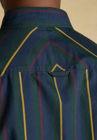 Detailný záber na zadnú stranu tmavomodrej košele so zelenými, žltými a červenými pruhmi, zobrazuje golier a malú textilnú slučku s etikettou "Joules".