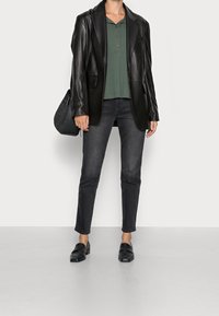 Blazer de cuero negro sobre un polo verde, combinado con jeans ajustados negros y mocasines. Se sostiene un bolso negro texturizado en una mano.