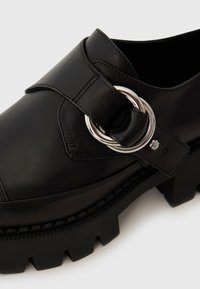 Black leather shoe with a thick, rugged sole. Features a wide strap and metallic circular hardware detail. Sleek design with smooth texture.
