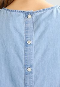 Haut en denim bleu clair avec un col arrondi et six boutons blancs dans le dos. Texture douce avec des détails de couture subtils.