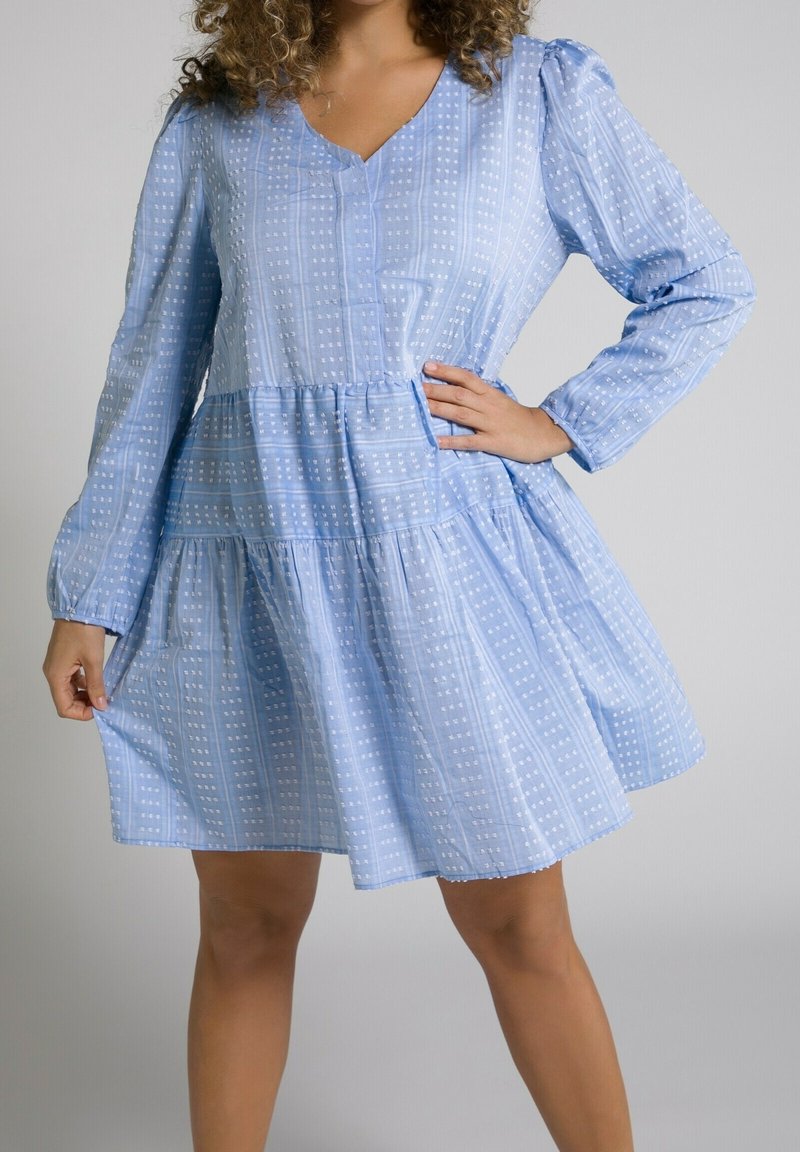 Femme portant une robe bleu clair à manches longues avec un motif et une jupe à volants, posant avec une main sur la hanche et l'autre tenant l'ourlet de la robe.