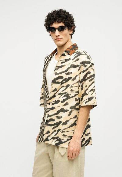 Άνδρας που φοράει κοντομάνικο πουκάμισο με ζωικά και καρό σχέδια, μπεζ παντελόνι, μαύρα γυαλιά ηλίου και λευκό εσώρουχο, στέκεται μπροστά σε λευκό φόντο.