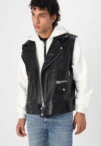 Black leather vest with zip closure, silver hardware, and logo detail. Worn over a white hoodie, paired with blue jeans.