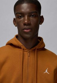 Felpa con cappuccio marrone con cerniera, realizzata in materiale morbido, caratterizzata da un logo bianco sul petto e cappuccio con coulisse. Texture liscia con una vestibilità aderente.