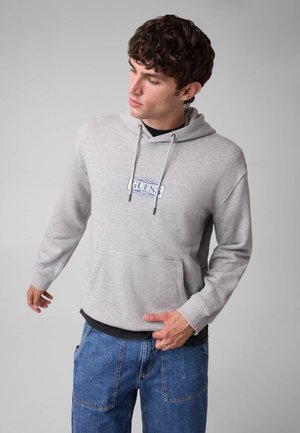 Giovane uomo che indossa una felpa Guess grigio chiaro e jeans blu guarda verso il basso con una mano vicino alla tasca, su uno sfondo grigio semplice.