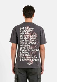 Grijze t-shirt met korte mouwen, voorzien van artistieke tekst en een eenvoudige lijntekening op de achterkant. De stof ziet er zacht en casual uit.