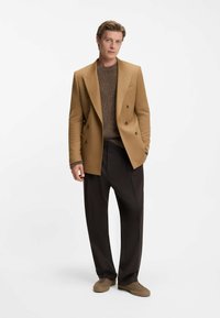 Blazer marrone doppiopetto su un maglione marrone a trama, abbinato a pantaloni scuri e scarpe marroni, il tutto su uno sfondo bianco.