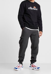 Sweat-shirt noir avec logo « ellesse », pantalon cargo gris avec poches latérales, et baskets blanches avec accents rouges et bleus.