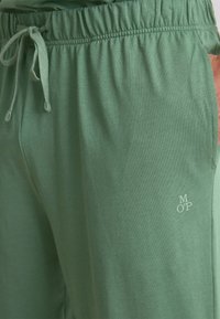 Zbliżenie na zielone spodnie z troczkiem w passie elastycznej i małym logo "MOP" na prawej udzie.