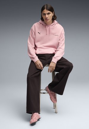 Jeune personne aux cheveux bruns mi-longs assise sur un tabouret, portant un sweat à capuche rose Puma Porsche, un pantalon large noir et des baskets roses.