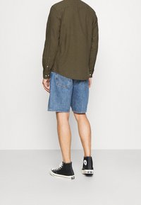 Camisa de manga larga verde oliva con botones, combinada con pantalones cortos de mezclilla azul claro. Zapatillas altas negras completan el look.