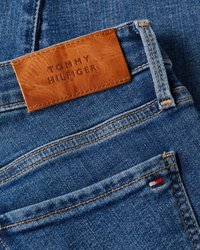 Džinsai su rudu odiniu žyme "TOMMY HILFIGER", mėlynos spalvos audinys su matomomis siūlėmis ir mažas vėliavos akcentas ant kišenės.