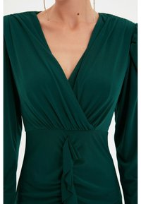 Femme portant une robe longue à manches longues de couleur vert profond avec un décolleté en V et un détail de tissu froncé à la taille.