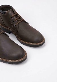 Par de botas de cuero marrón oscuro hasta el tobillo con cordones y suelas de goma negras sobre un fondo blanco.