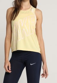 Top atlético amarillo sin mangas con un gran logo "NIKE" en rosa. Presenta un diseño sin mangas y un detalle de malla superpuesta. Combinado con leggins azul marino.