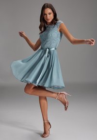 Une robe bleu clair avec un corsage en dentelle, des manches courtes et une jupe fluide. Elle présente une ceinture en satin et un nœud à l'arrière.