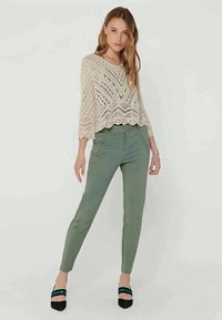 Beiger Strickpullover mit Lochmuster und gewelltem Saum, kombiniert mit schmal geschnittenen, olivgrünen Hosen und schwarzen High Heels mit türkisen Akzenten.