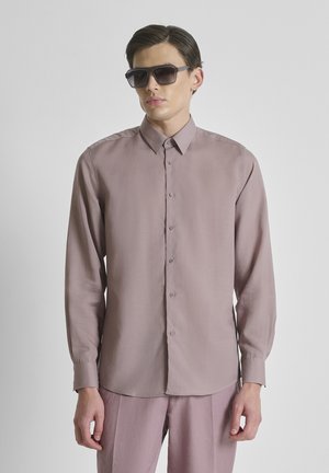 Mann mit zurückgestrichenen Haaren, der übergroße dunkle Sonnenbrillen, ein langärmeliges mauvefarbenes Hemd mit Knöpfen und passende Hose trägt, stehend vor weißem Hintergrund.